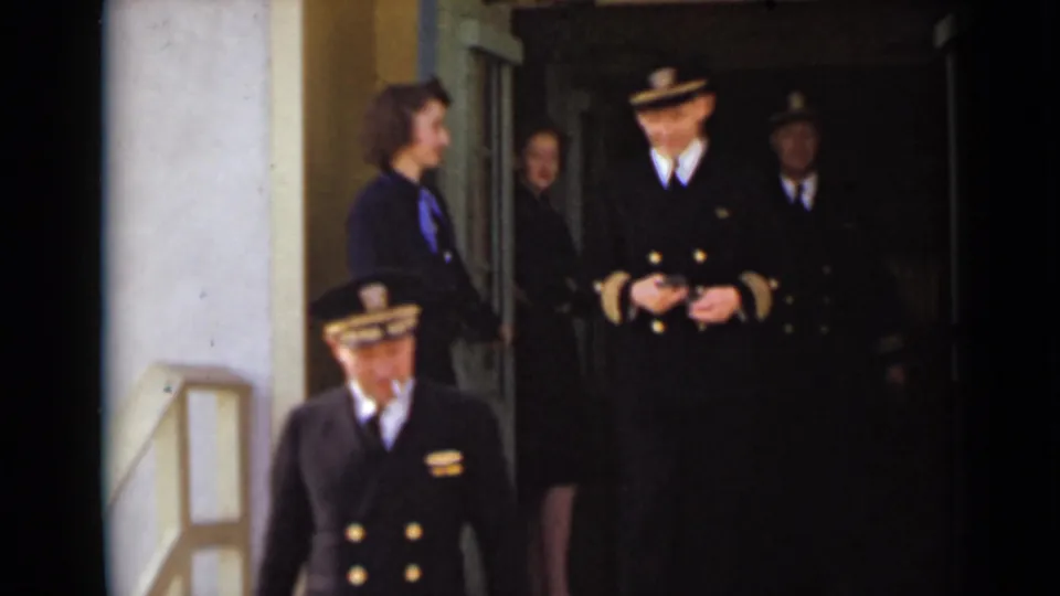 Uniformed adults stand and move at a New York building entrance, with a man in a peaked cap crossing the foreground.