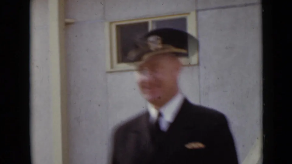 A uniformed man in a peaked cap passes close to the camera, blurred against a plain exterior wall and small window.