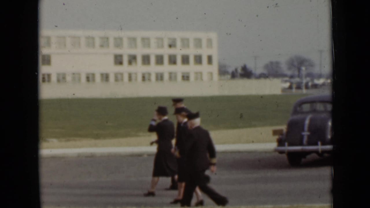 Uniformed Navy personnel walk along a paved area near a lawn and large building.