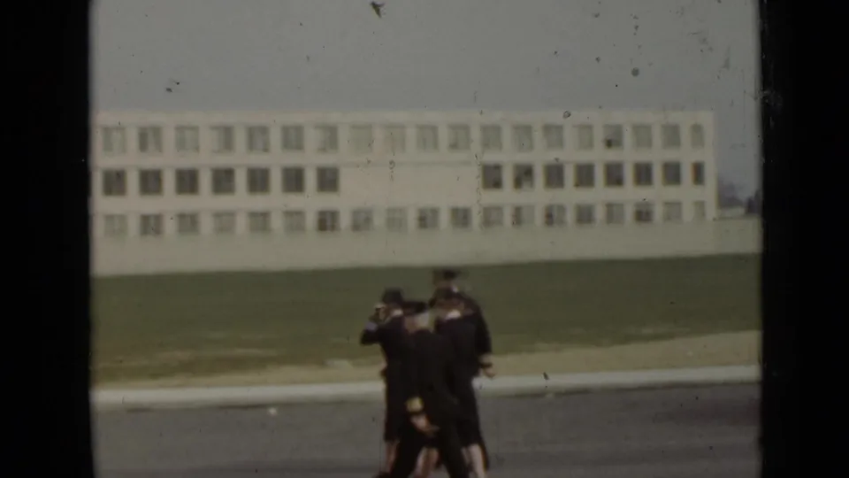 1947 New York 8mm frame showing four Navy personnel in formal uniforms walking together beside a lawn and institutional building.