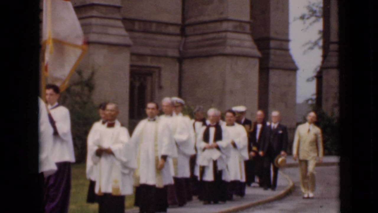 Clergy in white vestments and suited men walk past a stone church entrance.