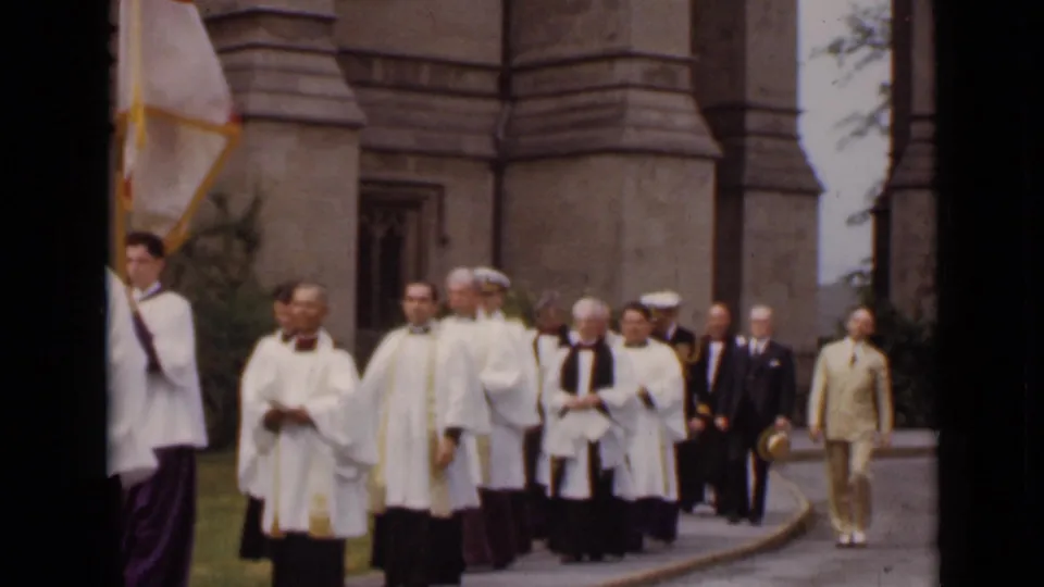 1947 New York home-movie frame showing clergy in white vestments and officials moving along a path outside a stone church.