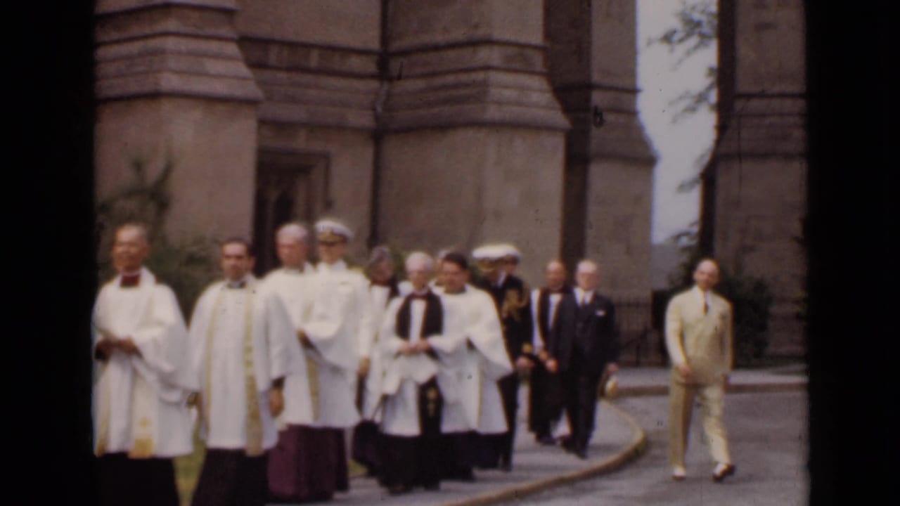 Religious procession continues beside a stone church with clergy, officers, and officials.