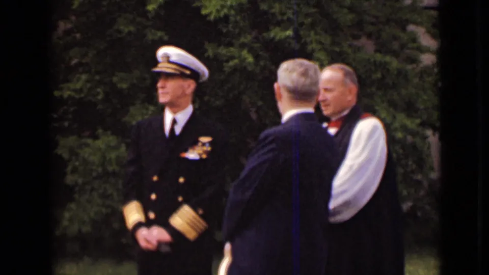 A naval officer in formal uniform stands while two men, one in dark clothing and one in white sleeves, converse on church grounds.