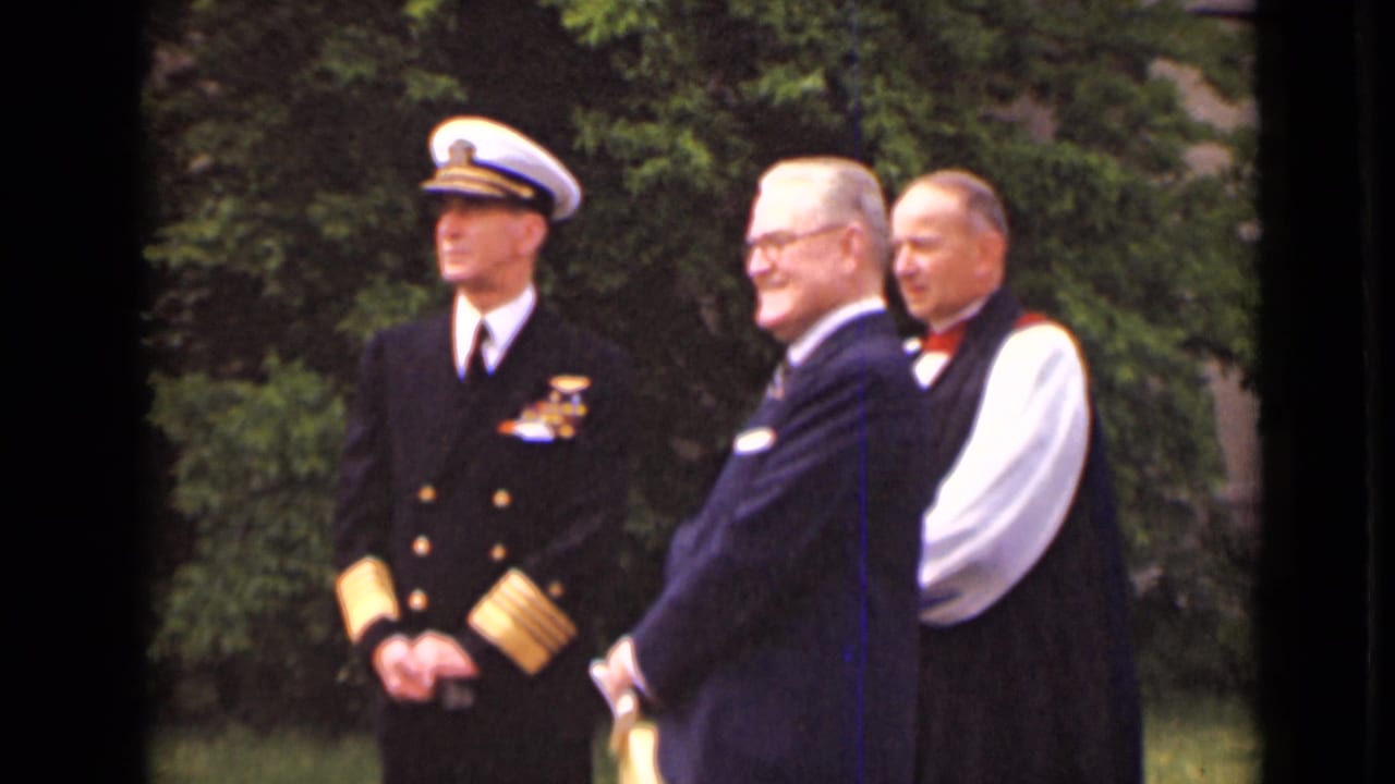 Naval officer in dress uniform stands beside two suited men outdoors.