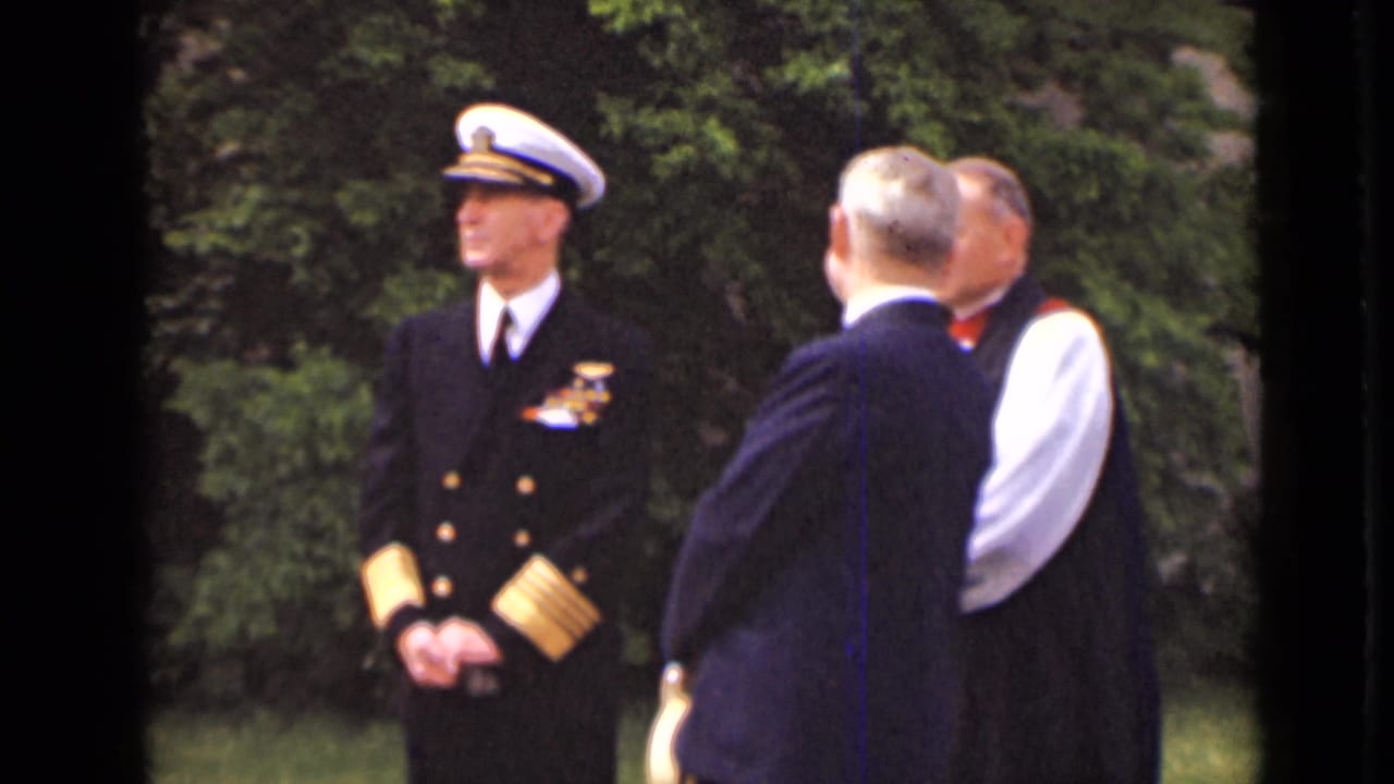 Officer in formal naval uniform faces suited men in a garden setting.