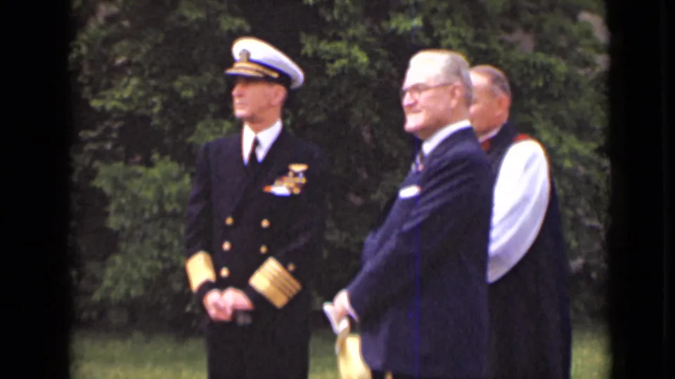 A 1947 New York frame shows the officer in profile with suited men beside him, filmed from a nearby home-movie viewpoint.