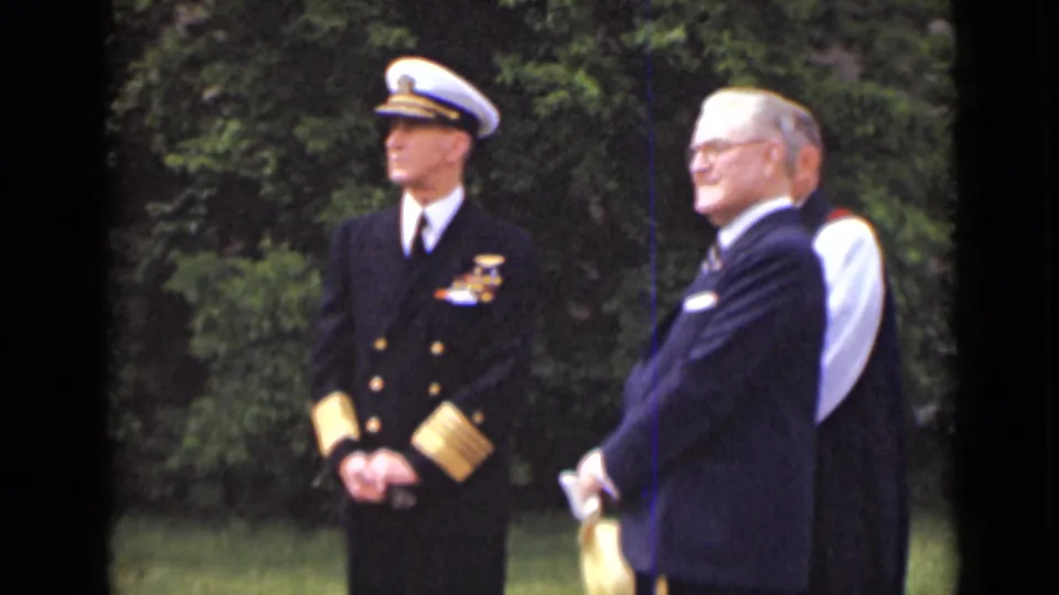 The final frame shows the naval officer standing formally while a suited man with glasses turns slightly toward the camera outdoors in New York.