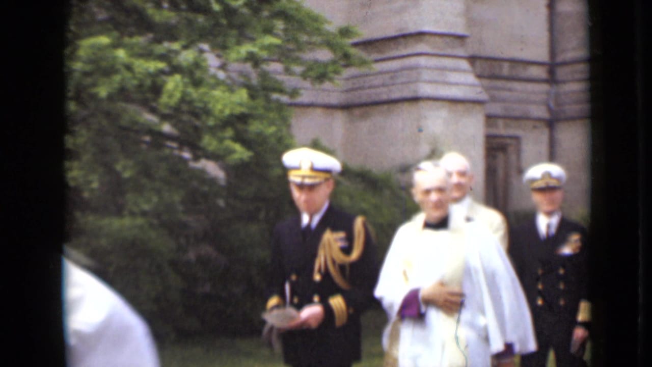Naval officer, clergyman, and suited men walking outdoors near trees and a stone building.