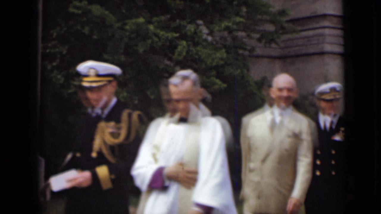 Clergyman in white vestments walks between a naval officer and men in light suits outdoors.