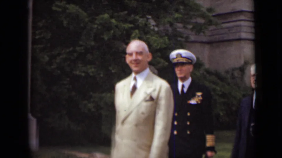 A bald man in a light suit faces the camera as a naval officer and other attendees remain near the stone building in 1947 New York footage.