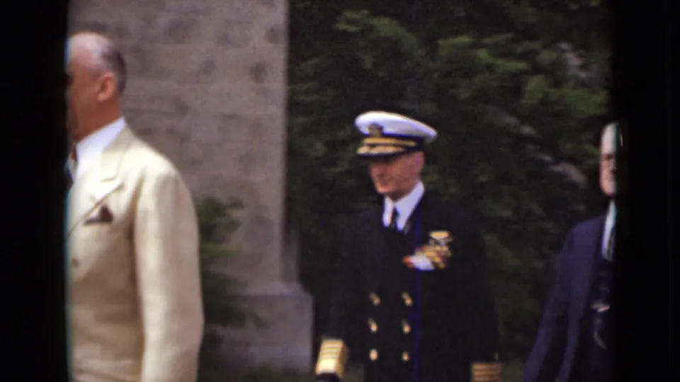 A man in a light suit crosses the foreground as a naval officer in dress uniform and other men remain visible in the 1947 New York scene.