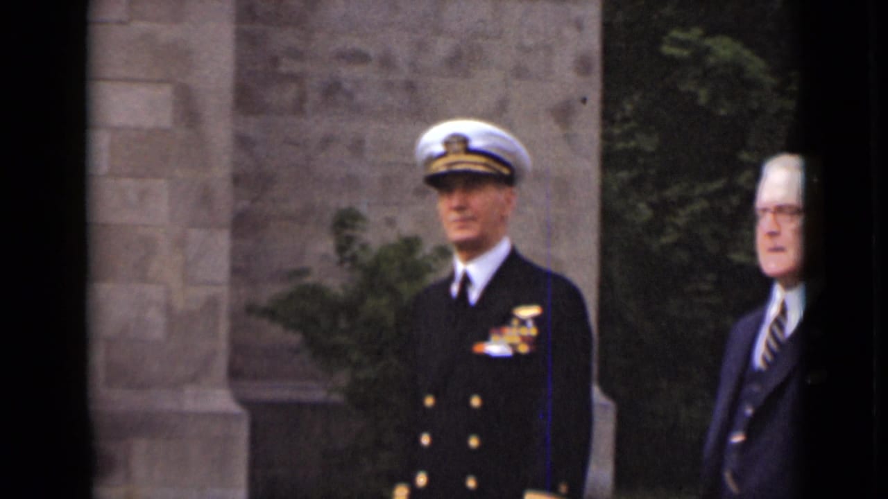 Naval officer and suited man standing near a stone building during a formal outdoor gathering.