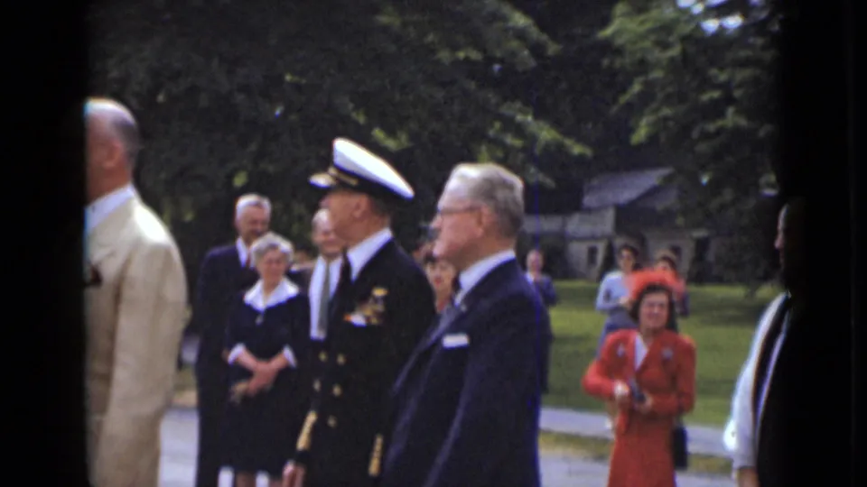 The naval officer and suited man are seen in profile while formally dressed spectators, including a woman in red, stand near the walkway.