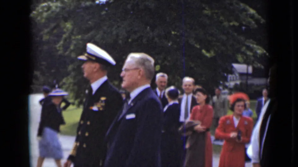 In this 1947 New York home-movie frame, a naval officer and suited man walk together as attendees and women in red stand behind them.