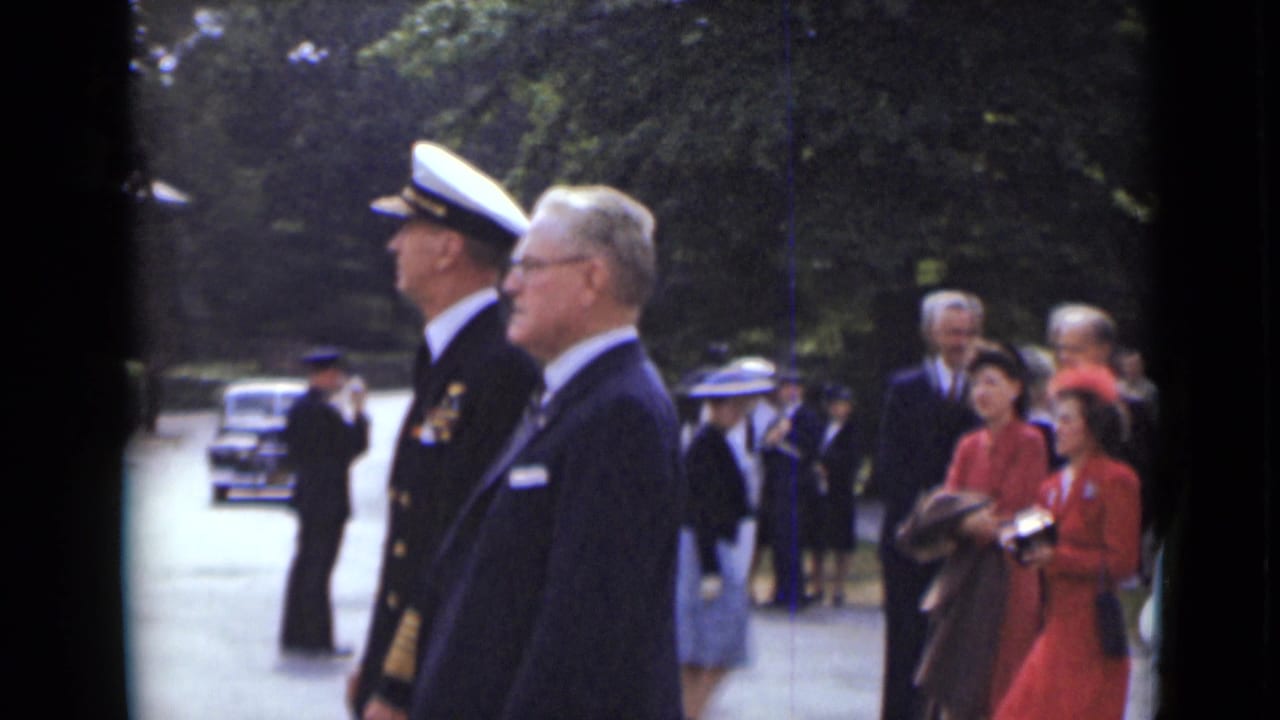Uniformed man and suited man pass a formal group on a tree-lined path.