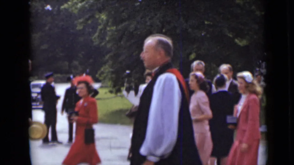 A clergyman in white robes and civilians in formal dresses and hats walk along a green outdoor path in New York archival footage.