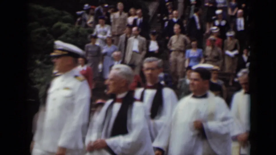 Clergy in white robes walk beside a uniformed man while a formally dressed crowd fills the steps behind them.