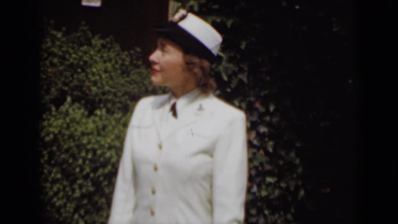 Woman in a white naval-style uniform and cap standing before dark green foliage.