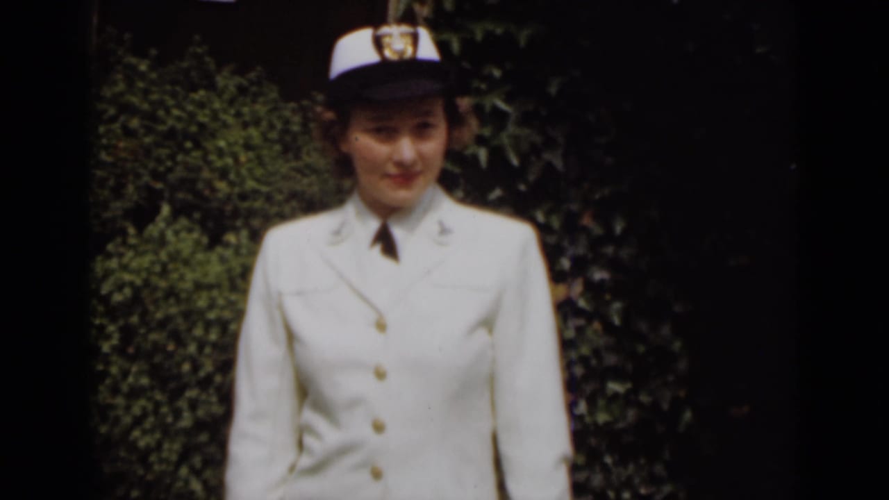 Woman in white uniform and cap looking toward the camera outdoors.