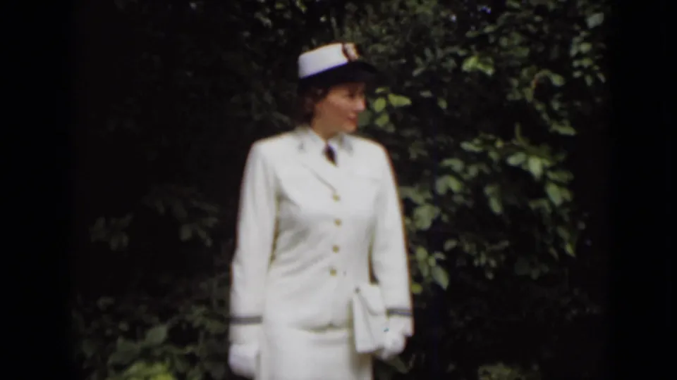 In 1947 New York footage, the woman stands in a white naval-style uniform, with gloves and a small handbag visible against dense greenery.