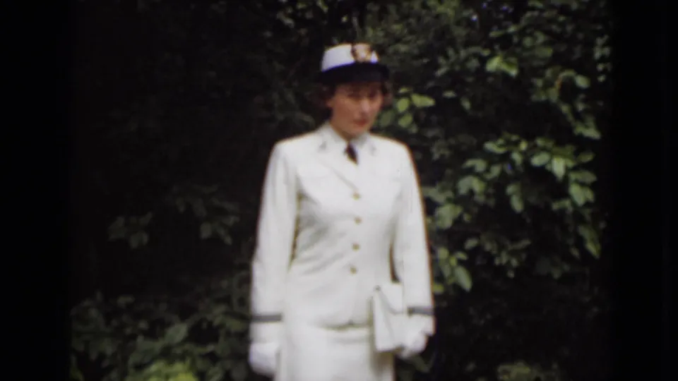 In 1947 New York footage, the uniformed woman stands near dark shrubs, her white jacket, gloves, cap, and handbag catching the light.