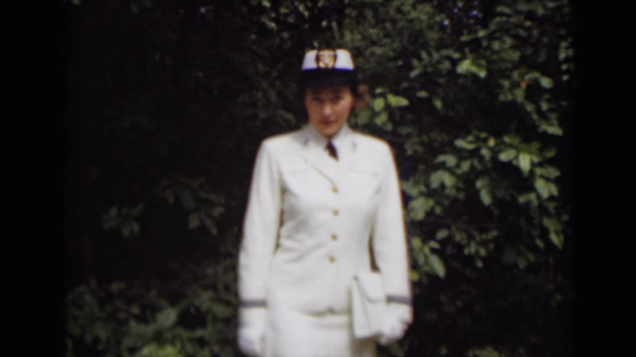 Woman in white uniform and cap standing before dark green foliage.