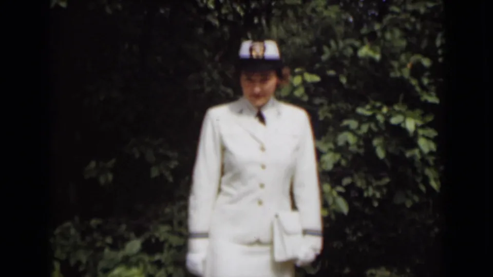 A woman in a white skirt uniform and cap stands outdoors against greenery in soft 8mm home-movie footage.