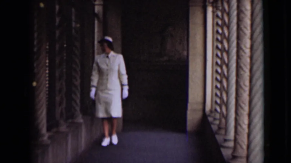 The woman in a white uniform walks along a shadowed colonnade with decorative columns and stone flooring.
