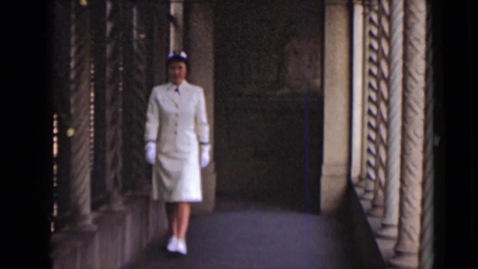 A woman in a white uniform faces the camera within a dim colonnade framed by ornate vertical columns.