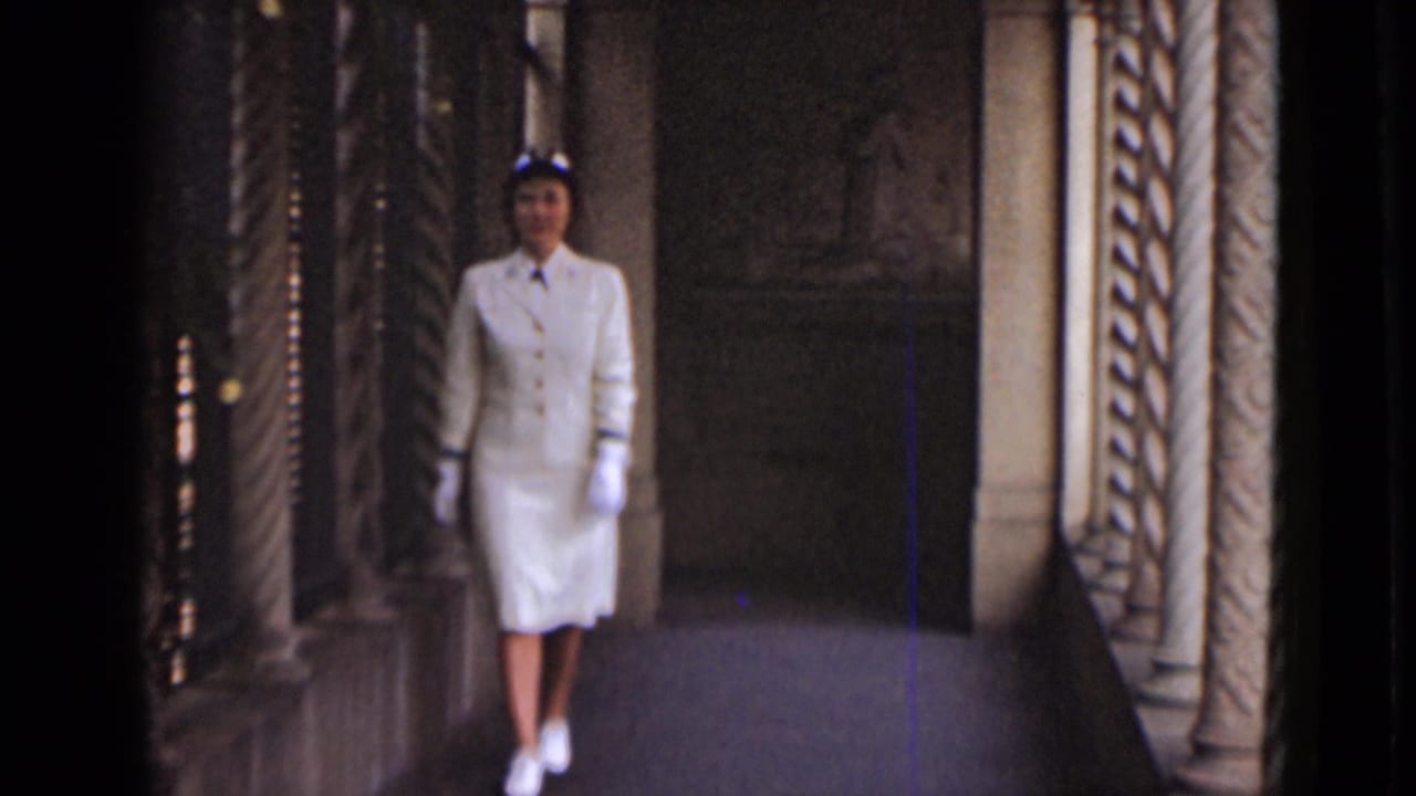 Woman in a white suit and hat walking down a stone corridor.