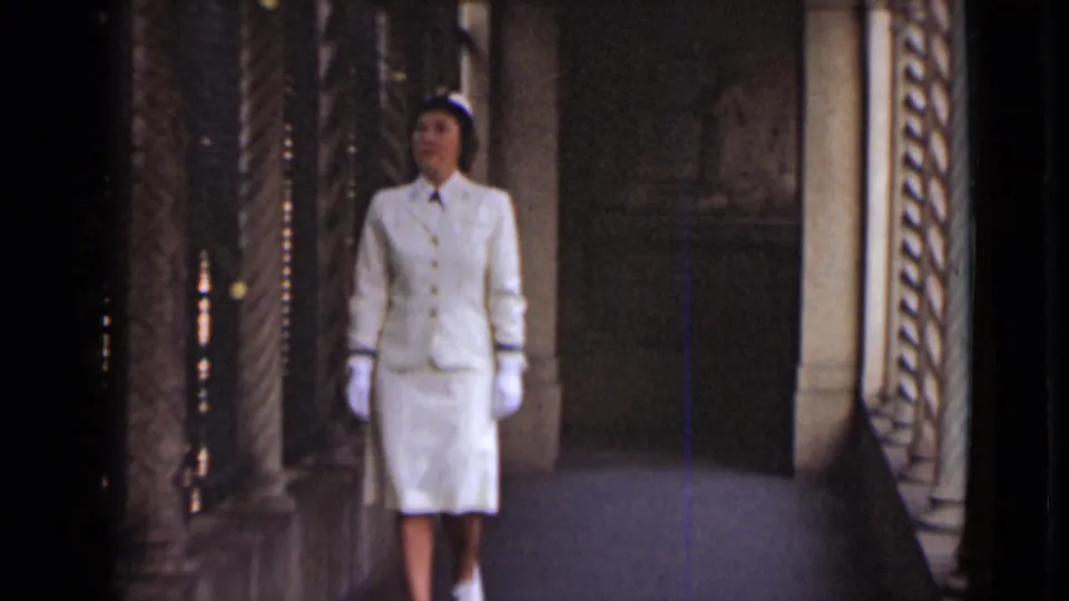 The woman in a white skirt suit and gloves faces forward in the New York corridor, framed by decorative stone columns and dim walls.