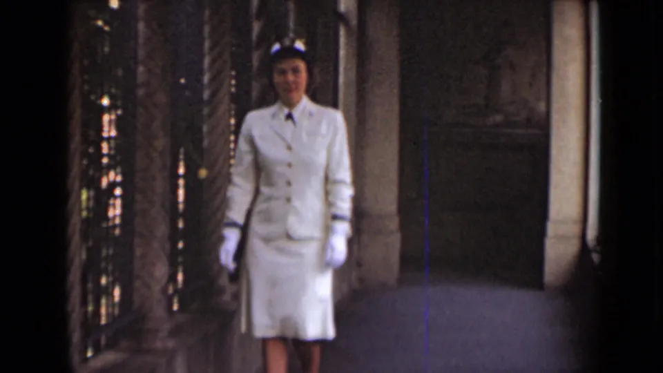 A closer frame shows the woman's white suit, gloves, hat, and shoes as she walks through the stone hallway in 1947 New York footage.