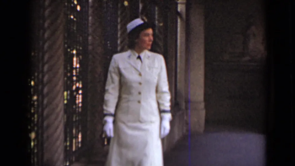 The woman turns to her right while standing in the ornate corridor, with her white gloves, skirt suit, and small hat visible.