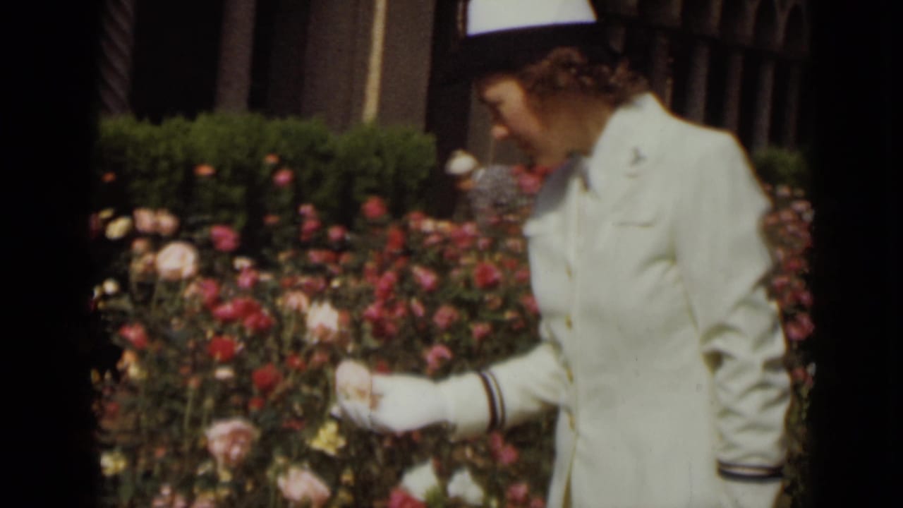 Woman in a white uniform bends toward rose blossoms in a garden.