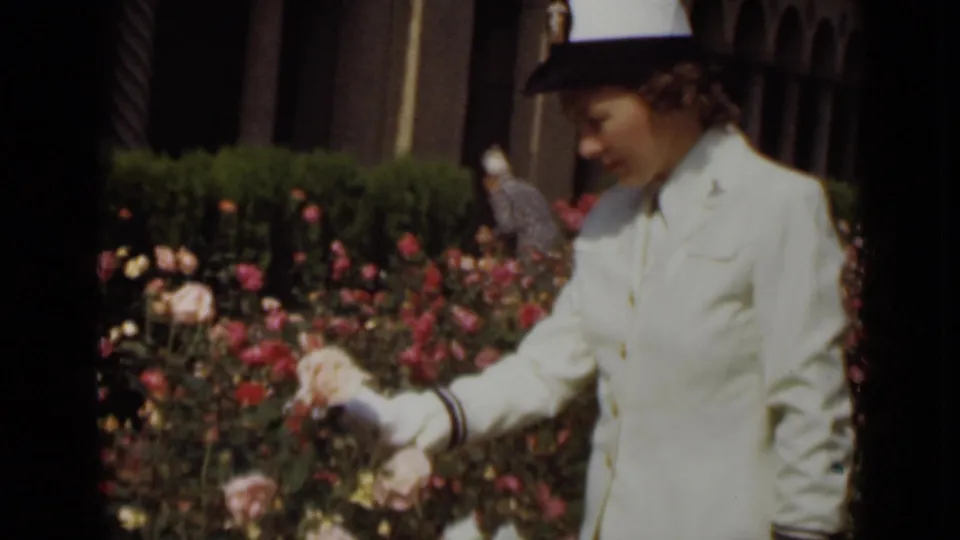 A gloved woman in a white uniform reaches toward pale rose blossoms, framed by flowering bushes and dark vertical garden elements.