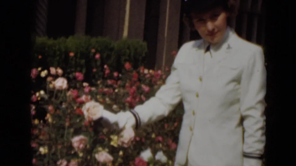 The uniformed woman looks down while touching a rose, with flowering bushes and greenery filling the garden background.