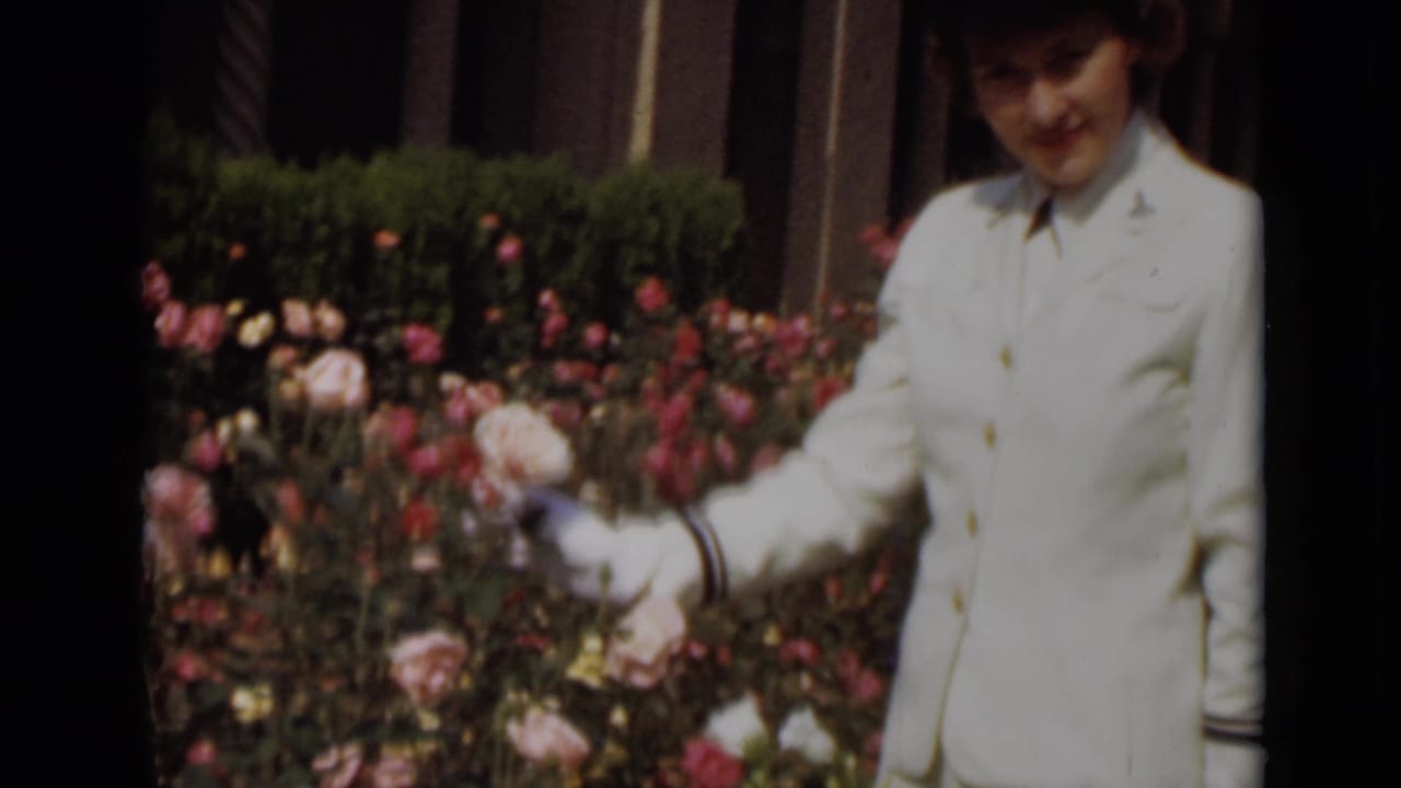 Young woman in a white uniform and gloves standing beside pink roses.