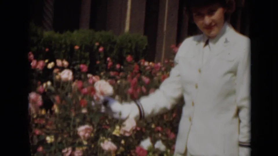 Young woman in a white uniform and gloves stands in a New York rose garden in 1947, holding a rose stem near the flowers.