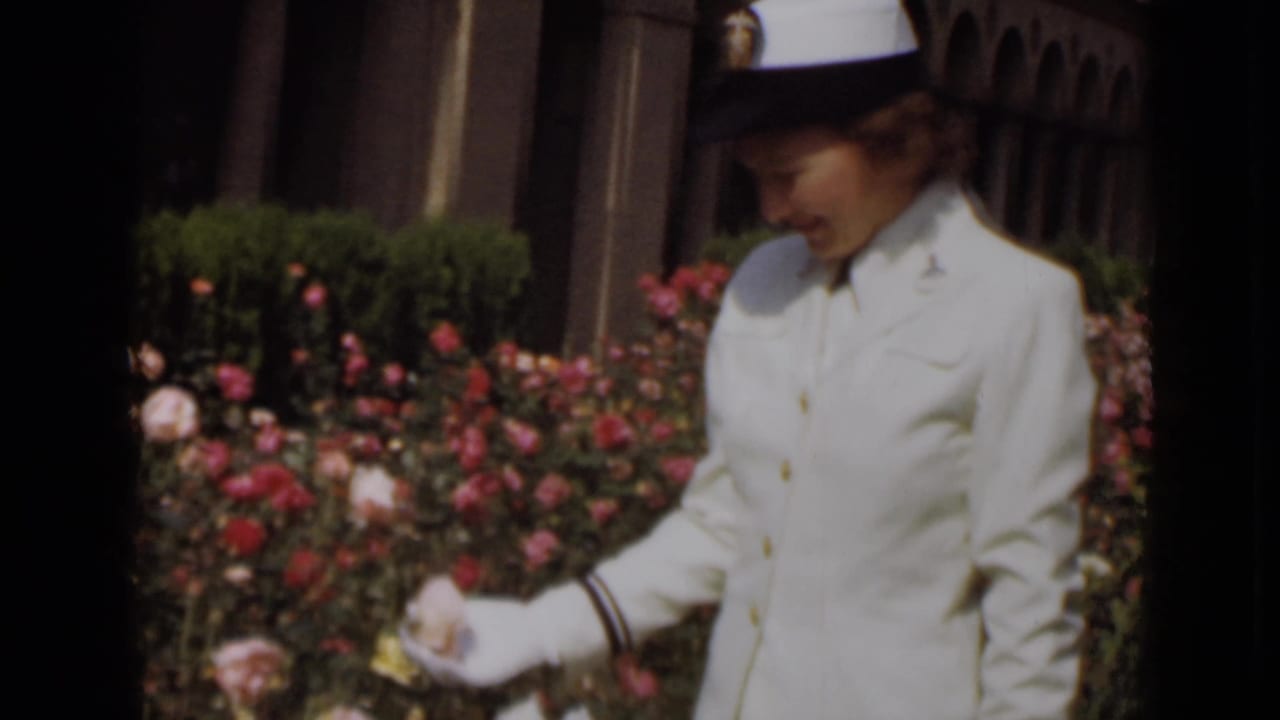 In 1947 New York footage, a woman in a white hat, uniform, and gloves looks down while touching roses in bright daylight.