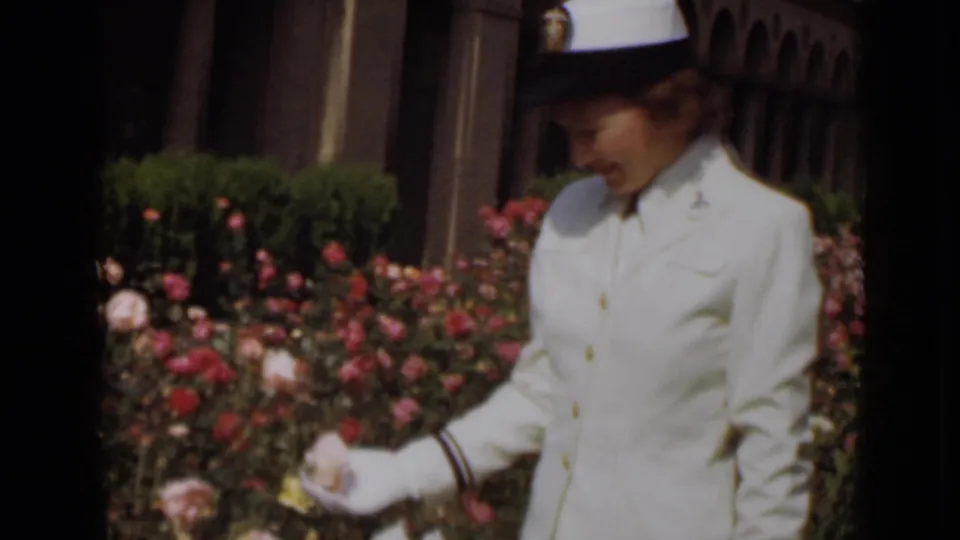 In 1947 New York footage, a woman in a white hat, uniform, and gloves looks down while touching roses in bright daylight.