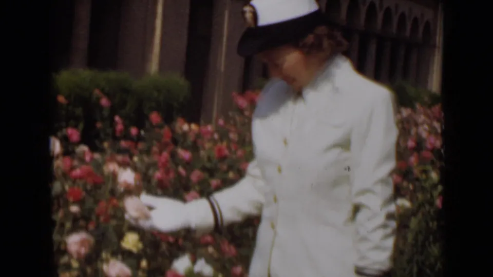 A uniformed woman in white gloves reaches toward pink roses in a 1947 New York 8mm home-movie frame.