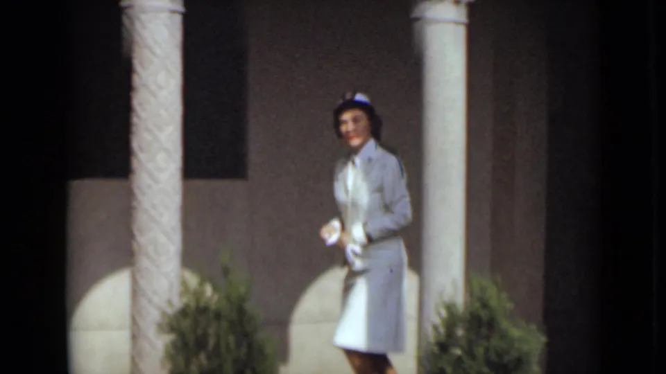 1947 New York frame showing an adult woman in a white dress, hat, and gloves walking beside pale columns.