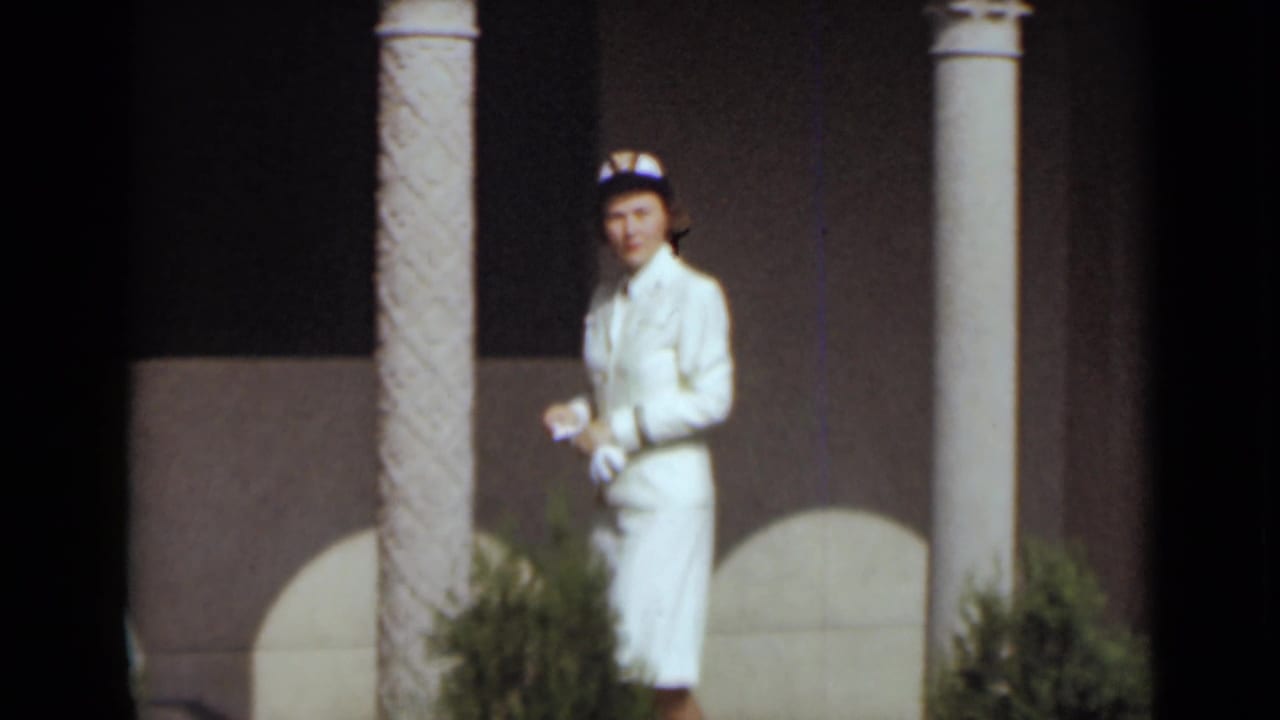 Woman in a white suit and hat standing beside columns in a formal garden.