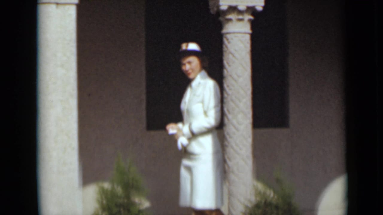 Woman in white standing near a column with shrubs in the foreground.
