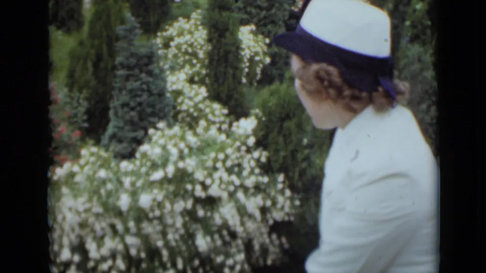 A closer 8mm frame shows the woman from behind and side as she looks across dense flowering shrubs and greenery.