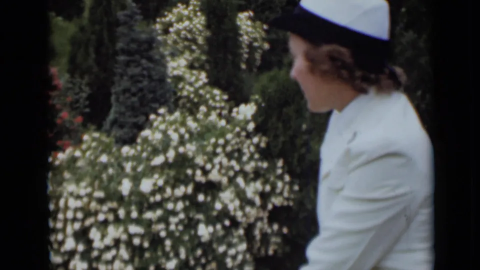 A side profile of the woman in a white suit and hat appears against a background of flowering shrubs and tall greenery.
