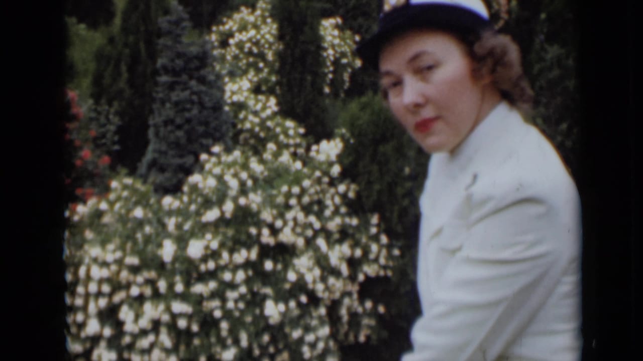 Woman in a white jacket and hat seated or standing beside flowering shrubs.