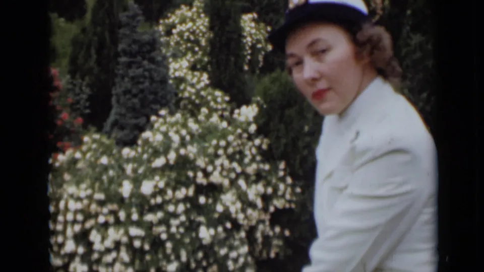 In 1947 New York home-movie footage, a woman in a white outfit and dark-brimmed hat looks down near a dense flowering bush.