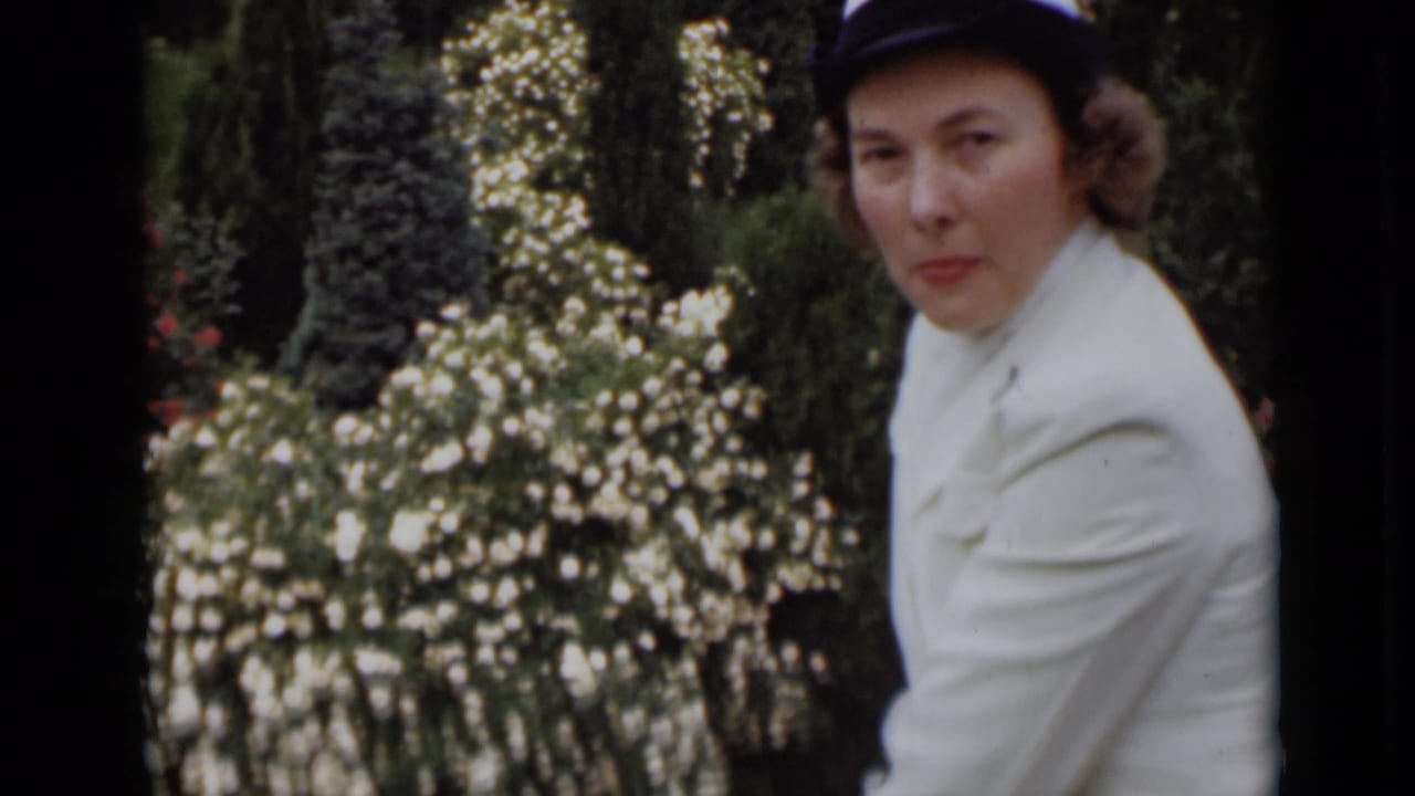 The woman in a white uniform-like outfit and hat faces the camera in a New York garden, with white blossoms filling the background.